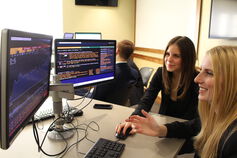 Two business students look at computer monitors showing stock market data.